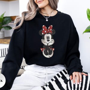 May include: Black sweatshirt featuring a distressed graphic of Minnie Mouse with a red polka dot bow. The sweatshirt has a crew neck and long sleeves. The word "BLACK" is printed on a white sticker in the lower left corner.