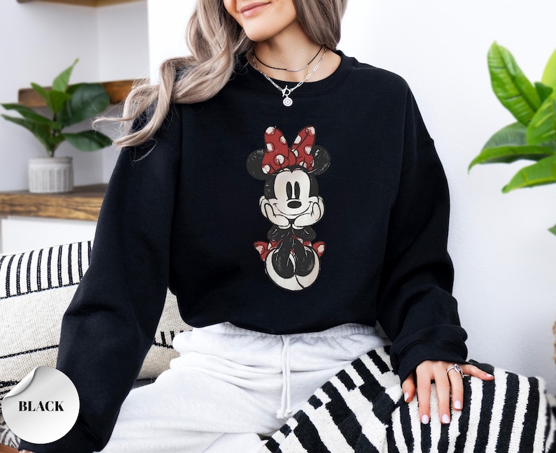 May include: Black sweatshirt featuring a distressed graphic of Minnie Mouse with a red polka dot bow. The sweatshirt has a crew neck and long sleeves. The word "BLACK" is printed on a white sticker in the lower left corner.