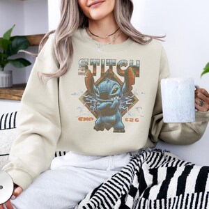 May include: A sand-colored crewneck sweatshirt featuring a graphic print of the character Stitch with arms crossed. The design includes the text STITCH above the character and EXP 626 below, set against a diamond-shaped background with lightning bolts.