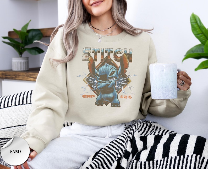 May include: A sand-colored crewneck sweatshirt featuring a graphic print of the character Stitch with arms crossed. The design includes the text STITCH above the character and EXP 626 below, set against a diamond-shaped background with lightning bolts.