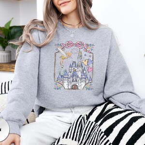 May include: A light grey sweatshirt featuring a colorful graphic of a fairytale castle, a fairy, and a pink bow. The design is framed with floral accents. The word "Party" is written above the castle. The sweatshirt is a comfortable, casual style.