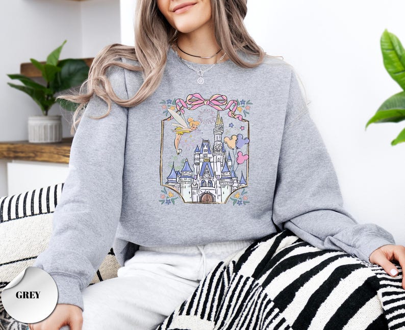 May include: A light grey sweatshirt featuring a colorful graphic of a fairytale castle, a fairy, and a pink bow. The design is framed with floral accents. The word "Party" is written above the castle. The sweatshirt is a comfortable, casual style.