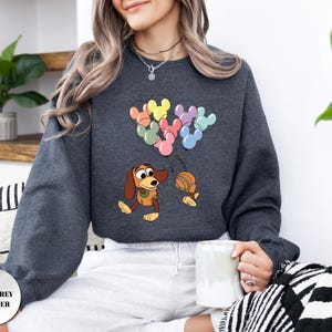 May include: Dark grey heather sweatshirt featuring a cartoon illustration of a dachshund dog holding a balloon string with Mickey Mouse-shaped balloons. The balloons are in various colors. The text "DARK GREY HEATHER" is in a circle at the bottom left.