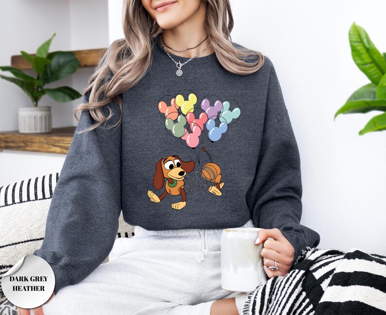 May include: Dark grey heather sweatshirt featuring a cartoon illustration of a dachshund dog holding a balloon string with Mickey Mouse-shaped balloons. The balloons are in various colors. The text "DARK GREY HEATHER" is in a circle at the bottom left.