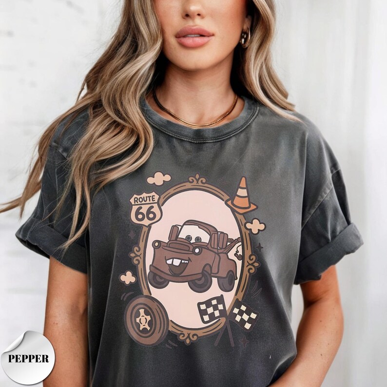 May include: A charcoal gray short-sleeve t-shirt featuring a graphic print of a brown tow truck character inside an oval frame. The design includes a Route 66 sign, a traffic cone, a tire, and two checkered flags. The word PEPPER is printed below.