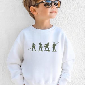 May include: White crewneck sweatshirt featuring a green silhouette of toy soldiers. The design includes four figures in various poses, suggesting a military theme. The sweatshirt is made of a soft material.