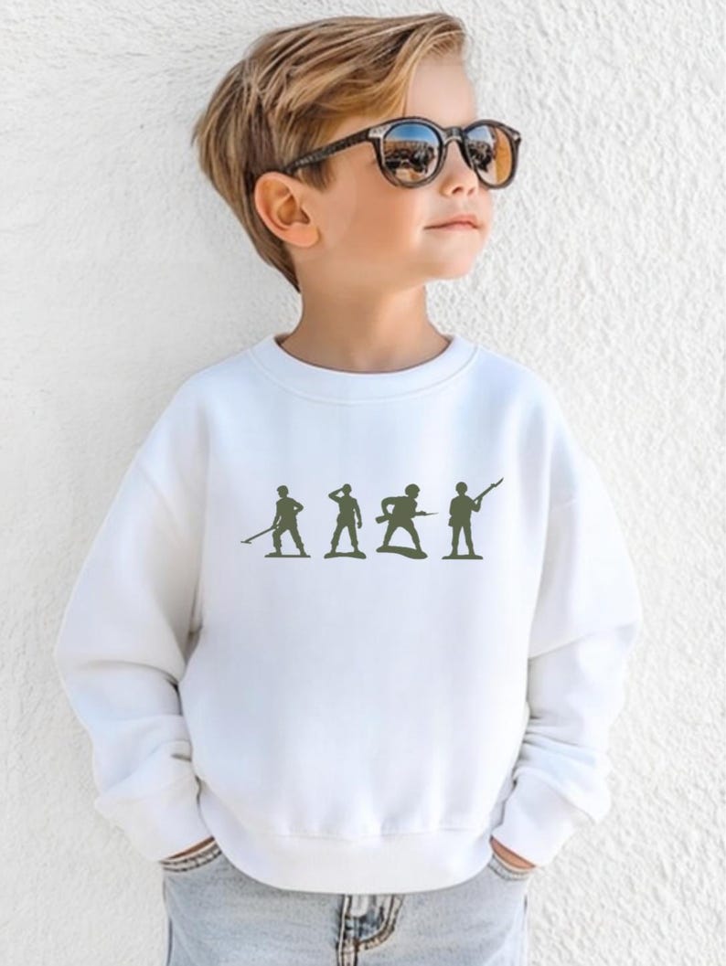 May include: White crewneck sweatshirt featuring a green silhouette of toy soldiers. The design includes four figures in various poses, suggesting a military theme. The sweatshirt is made of a soft material.