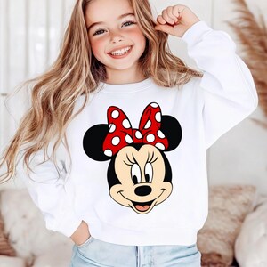 May include: White sweatshirt with a Minnie Mouse graphic. The design shows Minnie's face, including a red bow with white spots, black ears, and a smiling expression. A classic, comfortable style.