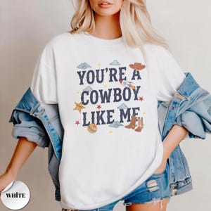May include: A white crewneck t-shirt featuring the dark blue text "YOU'RE A COWBOY LIKE ME" surrounded by colorful graphic illustrations including a rocket ship, a cowboy hat, a sheriff star, a ball, and small stars and clouds.