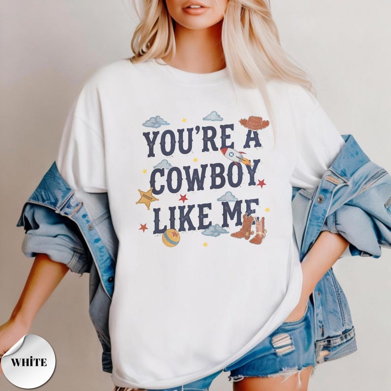May include: A white crewneck t-shirt featuring the dark blue text "YOU'RE A COWBOY LIKE ME" surrounded by colorful graphic illustrations including a rocket ship, a cowboy hat, a sheriff star, a ball, and small stars and clouds.