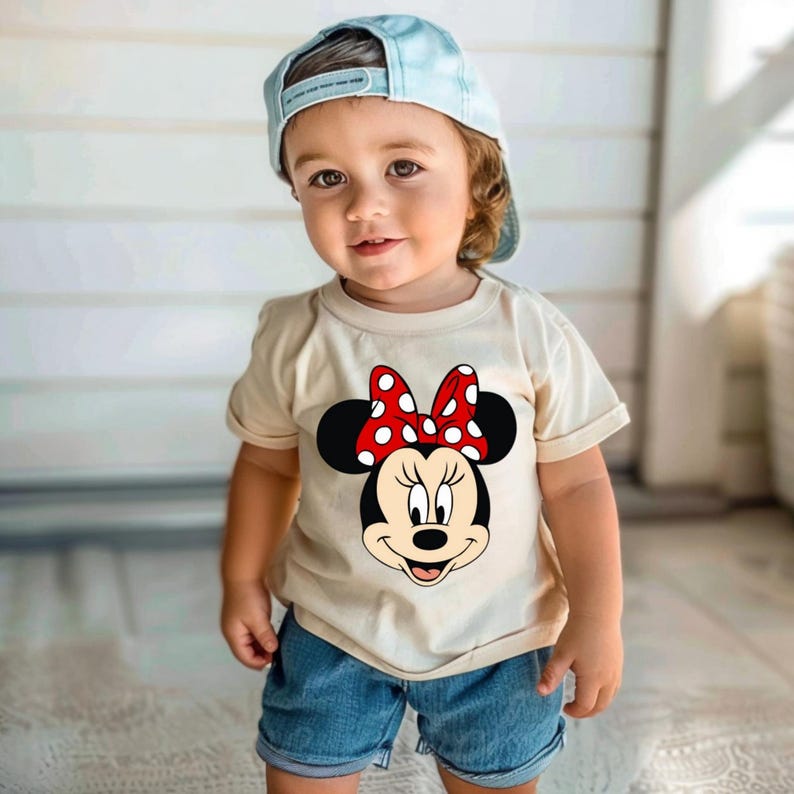 Puede incluir: Un ni&ntilde;o peque&ntilde;o con una camiseta beige claro con un gr&aacute;fico de Minnie Mouse. El gr&aacute;fico muestra la cara de Minnie con un lazo rojo con lunares blancos. El ni&ntilde;o tambi&eacute;n lleva pantalones cortos vaqueros azules y una gorra de b&eacute;isbol azul claro.