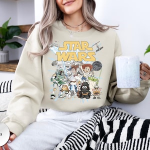 Puede incluir: Una sudadera de cuello redondo color arena con un estampado gr&aacute;fico de personajes de Star Wars reimaginados como figuras de Toy Story. El dise&ntilde;o incluye el logotipo amarillo de Star Wars sobre ilustraciones de personajes como Woody, Buzz y Jessie.