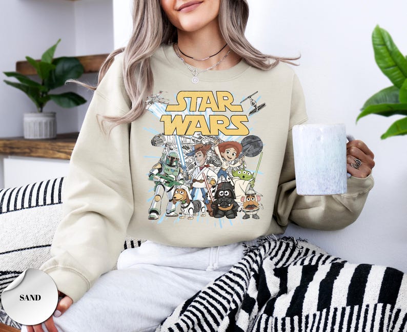 Puede incluir: Una sudadera de cuello redondo color arena con un estampado gr&aacute;fico de personajes de Star Wars reimaginados como figuras de Toy Story. El dise&ntilde;o incluye el logotipo amarillo de Star Wars sobre ilustraciones de personajes como Woody, Buzz y Jessie.
