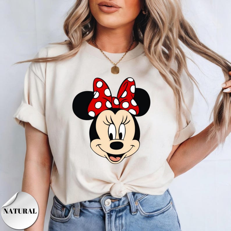 Puede incluir: Camiseta color crema con un gr&aacute;fico de la cara de Minnie Mouse. La cara de Minnie es negra con un lazo rojo y blanco de lunares. La palabra "NATURAL" est&aacute; impresa en una etiqueta blanca en la esquina inferior izquierda.