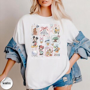 K&ouml;nnte beinhalten: Wei&szlig;es T-Shirt mit einer farbenfrohen Collage von Disney-Figuren und Wahrzeichen. Das Design umfasst Micky Maus, Minnie Maus, Donald Duck und Chip und Chap, mit dem Text "Walt Disney World" und "2026".