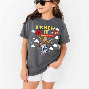 Puede incluir: Una camiseta gris carb&oacute;n con un gr&aacute;fico de una persona de pelo largo tocando la guitarra, con una camisa de cuadros amarilla y pantalones de campana con estampado de vaca. El texto dice I KNEW IT y I KNEW YOU, junto a notas musicales y estrellas.