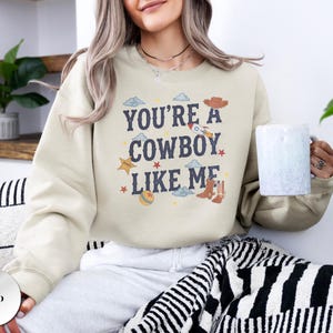 May include: A sand-colored crewneck sweatshirt featuring the dark blue text "YOU'RE A COWBOY LIKE ME" centered on the chest. The design includes small graphic illustrations of a cowboy hat, a rocket, a star, a ball, and cowboy boots.