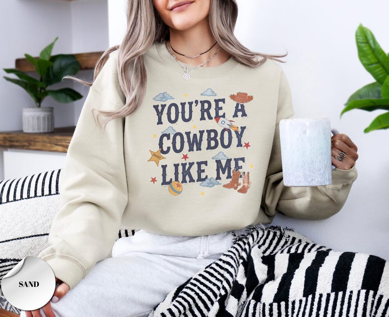 May include: A sand-colored crewneck sweatshirt featuring the dark blue text "YOU'RE A COWBOY LIKE ME" centered on the chest. The design includes small graphic illustrations of a cowboy hat, a rocket, a star, a ball, and cowboy boots.