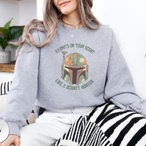 Puede incluir: Una sudadera gris jaspeado de cuello redondo con un gr&aacute;fico de un casco de cazarrecompensas en tonos verde, naranja y marr&oacute;n. El texto "KARMA'S ON YOUR SCENT LIKE A BOUNTY HUNTER" est&aacute; impreso en una fuente arqueada verde oscuro alrededor.