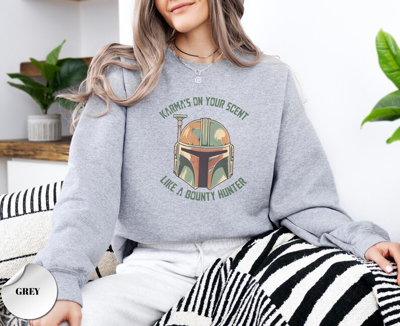 Puede incluir: Una sudadera gris jaspeado de cuello redondo con un gr&aacute;fico de un casco de cazarrecompensas en tonos verde, naranja y marr&oacute;n. El texto "KARMA'S ON YOUR SCENT LIKE A BOUNTY HUNTER" est&aacute; impreso en una fuente arqueada verde oscuro alrededor.