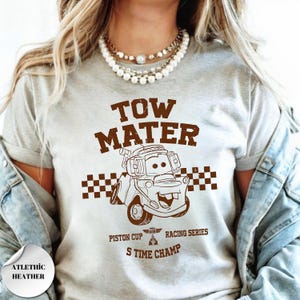 May include: A light gray t-shirt featuring a brown graphic of the character Tow Mater from the movie Cars. The design includes a checkered flag pattern, the text TOW MATER, PISTON CUP RACING SERIES, and S TIME CHAMP, with a small trophy icon.