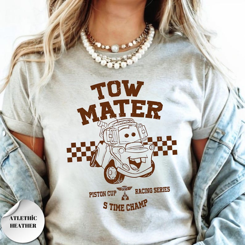 May include: A light gray t-shirt featuring a brown graphic of the character Tow Mater from the movie Cars. The design includes a checkered flag pattern, the text TOW MATER, PISTON CUP RACING SERIES, and S TIME CHAMP, with a small trophy icon.