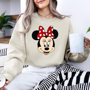 May include: A sand-coloured sweatshirt with a large graphic of Minnie Mouse's face. Minnie's face is black and white, with a red bow with white polka dots. The sweatshirt is a casual style.