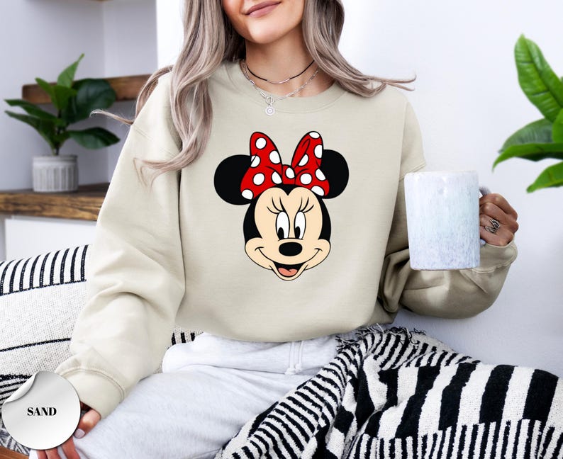 May include: A sand-coloured sweatshirt with a large graphic of Minnie Mouse's face. Minnie's face is black and white, with a red bow with white polka dots. The sweatshirt is a casual style.