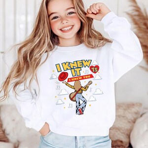 May include: A white long-sleeve sweatshirt featuring a graphic print of a person with long hair playing a guitar, wearing cow-print trousers and a red hat. The text reads I KNEW IT I KNEW YOU with musical notes, stars, clouds, and a red heart with TS.