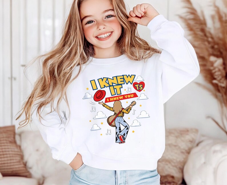 May include: A white long-sleeve sweatshirt featuring a graphic print of a person with long hair playing a guitar, wearing cow-print trousers and a red hat. The text reads I KNEW IT I KNEW YOU with musical notes, stars, clouds, and a red heart with TS.