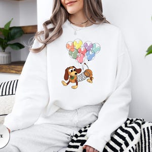 May include: White sweatshirt featuring a cartoon illustration of a dachshund dog holding colorful Mickey Mouse-shaped balloons. The dog has a spring-like body and is wearing a green collar. The word "WHITE" is printed on a white circle.