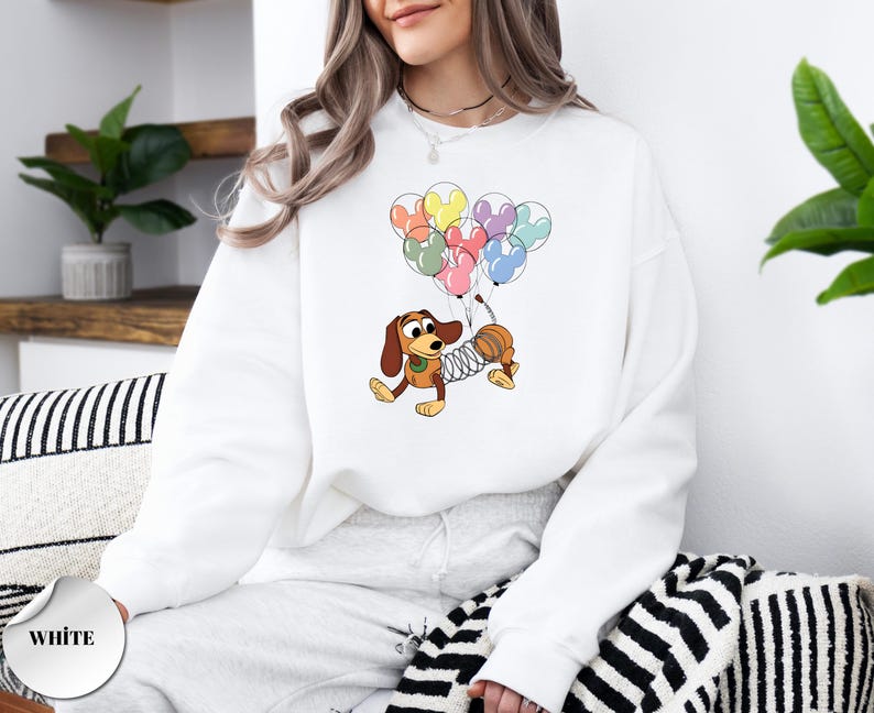 May include: White sweatshirt featuring a cartoon illustration of a dachshund dog holding colorful Mickey Mouse-shaped balloons. The dog has a spring-like body and is wearing a green collar. The word "WHITE" is printed on a white circle.