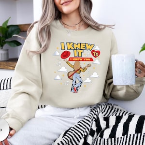 May include: A sand-coloured crewneck sweatshirt featuring a graphic print of a person with long hair playing a guitar, wearing a yellow shirt and blue bell-bottom trousers. The text reads I KNEW IT I KNEW YOU with a red hat, a red heart, and clouds.
