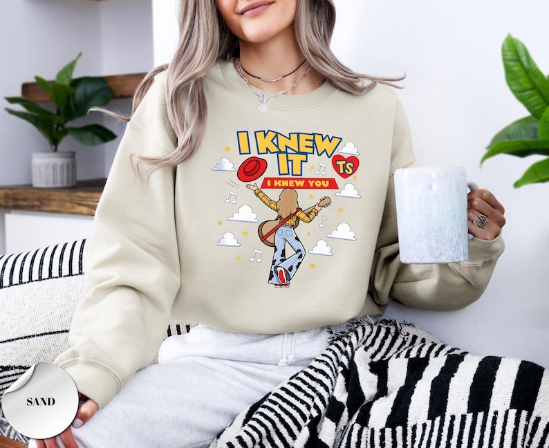 May include: A sand-coloured crewneck sweatshirt featuring a graphic print of a person with long hair playing a guitar, wearing a yellow shirt and blue bell-bottom trousers. The text reads I KNEW IT I KNEW YOU with a red hat, a red heart, and clouds.