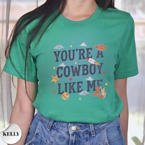 May include: A green short-sleeve t-shirt featuring the dark blue text "YOU'RE A COWBOY LIKE ME" centered on the chest. The design includes small graphic illustrations of a brown cowboy hat, a rocket, a sheriff star, a ball, and cowboy boots.