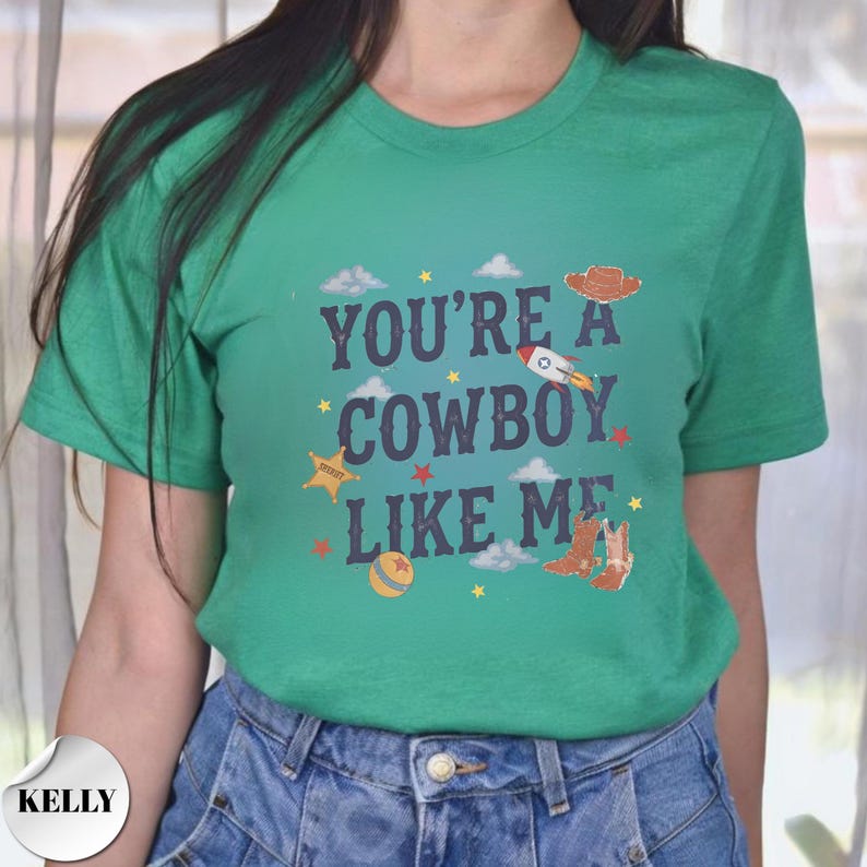 May include: A green short-sleeve t-shirt featuring the dark blue text "YOU'RE A COWBOY LIKE ME" centered on the chest. The design includes small graphic illustrations of a brown cowboy hat, a rocket, a sheriff star, a ball, and cowboy boots.