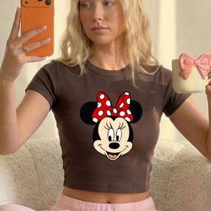 Puede incluir: Un top corto marr&oacute;n con un dise&ntilde;o de Minnie Mouse. El dise&ntilde;o incluye la cara de Minnie, orejas negras y un lazo rojo con lunares blancos. El top tiene mangas cortas y cuello redondo. La imagen tambi&eacute;n muestra una taza de color claro con un lazo rosa.