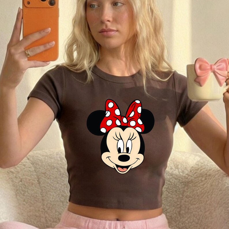 Puede incluir: Un top corto marr&oacute;n con un dise&ntilde;o de Minnie Mouse. El dise&ntilde;o incluye la cara de Minnie, orejas negras y un lazo rojo con lunares blancos. El top tiene mangas cortas y cuello redondo. La imagen tambi&eacute;n muestra una taza de color claro con un lazo rosa.