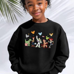 Op de afbeelding: Een zwart sweatshirt met ronde hals en een kleurrijke grafische print van Toy Story-personages, waaronder Rex, Hamm, Woody, Buzz Lightyear, Bullseye en Jessie, geaccentueerd door kleine zwevende Mickey Mouse-oorpictogrammen in diverse patronen.