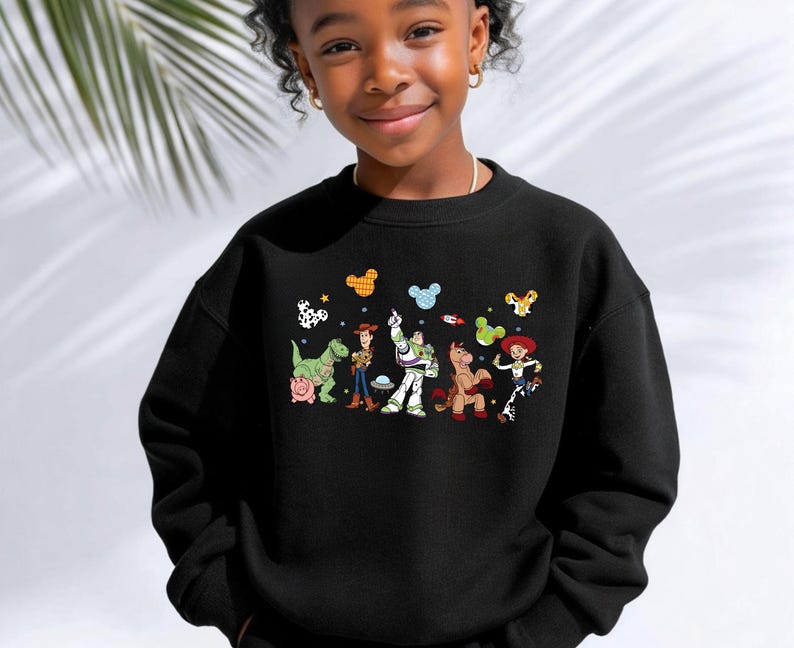 Op de afbeelding: Een zwart sweatshirt met ronde hals en een kleurrijke grafische print van Toy Story-personages, waaronder Rex, Hamm, Woody, Buzz Lightyear, Bullseye en Jessie, geaccentueerd door kleine zwevende Mickey Mouse-oorpictogrammen in diverse patronen.