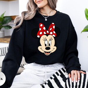 May include: Black sweatshirt with a Minnie Mouse design. The graphic depicts Minnie's face, complete with a red bow adorned with white polka dots. The word "BLACK" is printed on a white circle in the lower left corner. A casual style sweatshirt.
