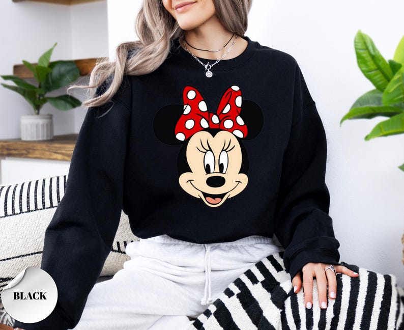 May include: Black sweatshirt with a Minnie Mouse design. The graphic depicts Minnie's face, complete with a red bow adorned with white polka dots. The word "BLACK" is printed on a white circle in the lower left corner. A casual style sweatshirt.