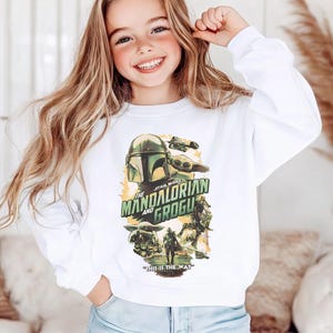 Puede incluir: Una sudadera blanca de manga larga con un estampado gr&aacute;fico de The Mandalorian y Grogu. El dise&ntilde;o incluye el texto "The Mandalorian and Grogu" y "This is the way" con varias ilustraciones de personajes en tonos verdes, marrones y amarillos.