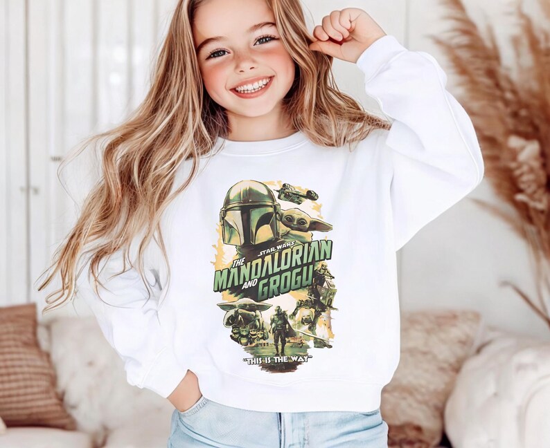 Puede incluir: Una sudadera blanca de manga larga con un estampado gr&aacute;fico de The Mandalorian y Grogu. El dise&ntilde;o incluye el texto "The Mandalorian and Grogu" y "This is the way" con varias ilustraciones de personajes en tonos verdes, marrones y amarillos.