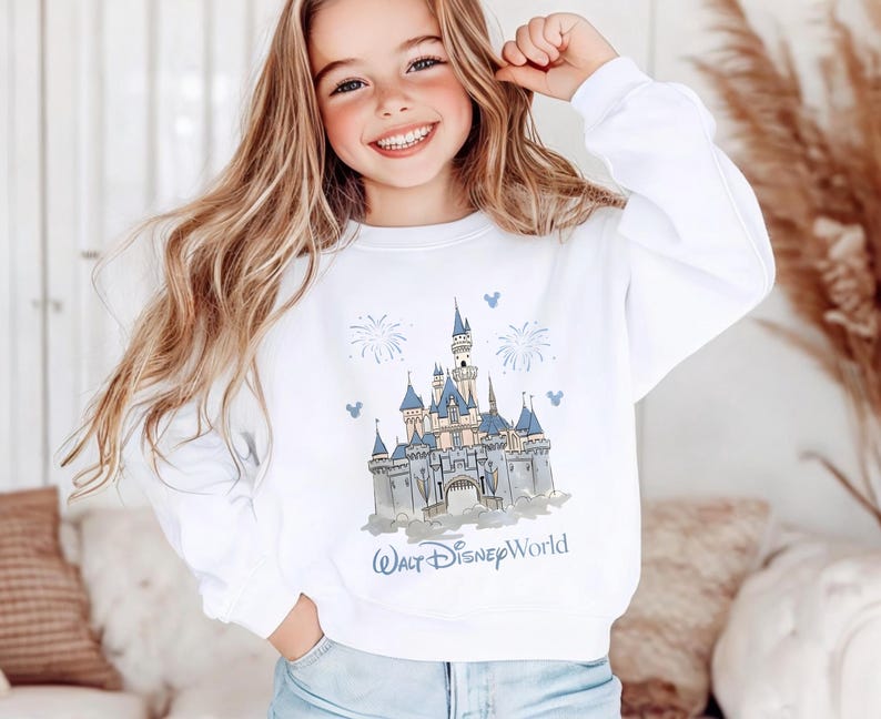 May include: A white long-sleeve sweatshirt featuring a graphic illustration of a castle with blue roofs and spires, surrounded by fireworks and small mouse-ear icons. The text Walt Disney World is printed in blue below the castle illustration.
