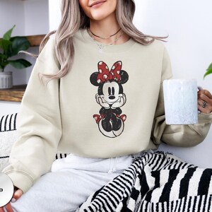 May include: Sand-colored sweatshirt featuring a distressed graphic of Minnie Mouse. Minnie is depicted with a red bow with white polka dots, a black dress, and red shoes. The sweatshirt is a crew neck style.
