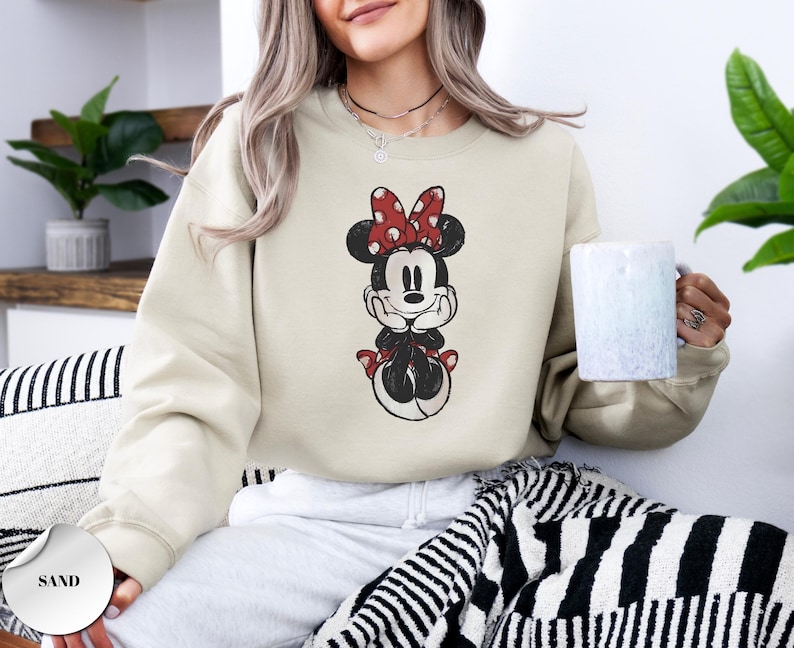 May include: Sand-colored sweatshirt featuring a distressed graphic of Minnie Mouse. Minnie is depicted with a red bow with white polka dots, a black dress, and red shoes. The sweatshirt is a crew neck style.