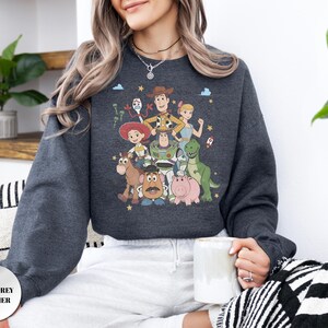 May include: A dark grey heather long-sleeve sweatshirt featuring a colorful graphic print of various animated characters including a cowboy, a space ranger, a cowgirl, a dinosaur, a pig, a potato-shaped figure, and a small white toy.