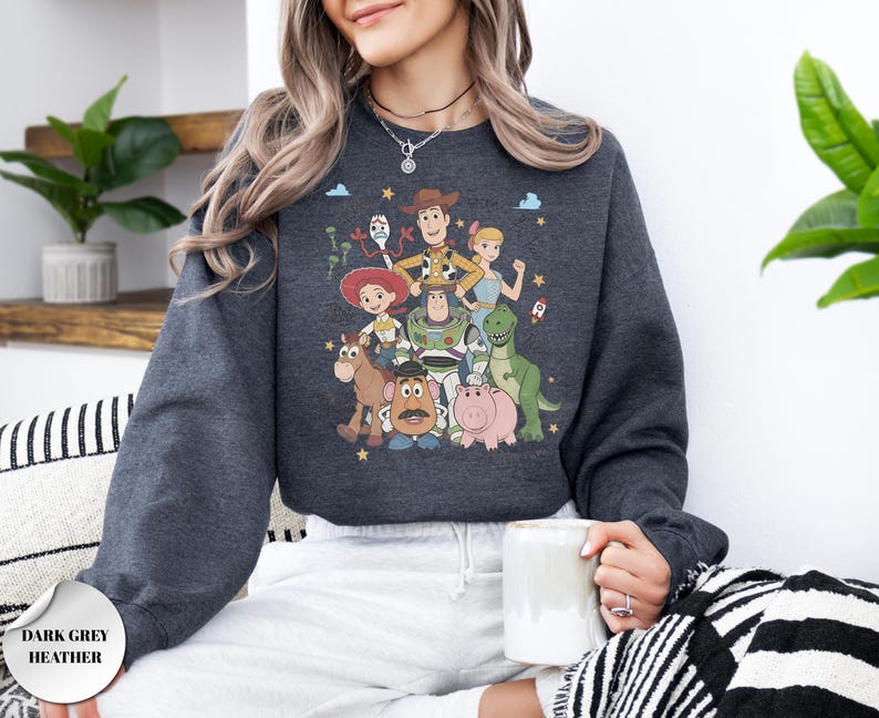 May include: A dark grey heather long-sleeve sweatshirt featuring a colorful graphic print of various animated characters including a cowboy, a space ranger, a cowgirl, a dinosaur, a pig, a potato-shaped figure, and a small white toy.