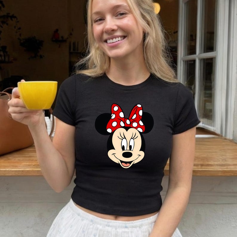 Puede incluir: Camiseta negra de cuello redondo con un gr&aacute;fico de Minnie Mouse. El gr&aacute;fico muestra la cara de Minnie con un lazo rojo con lunares blancos. La camiseta es de estilo ajustado.